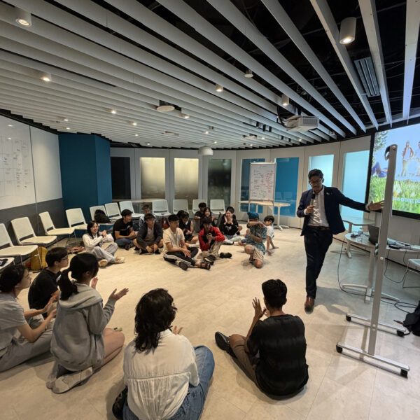A facilitator leads a workshop for students sitting in a circle, gesturing toward a screen prompt to plan a class picnic in Youth leadership and Communication Camp