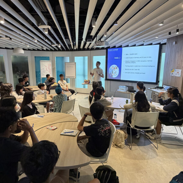 A presenter stands before a group of students seated at round tables during a youth leadership workshop