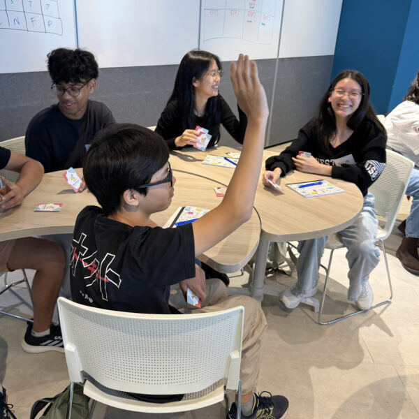 Students smiling and participating in a workshop activity at a table, with one student raising their hand
