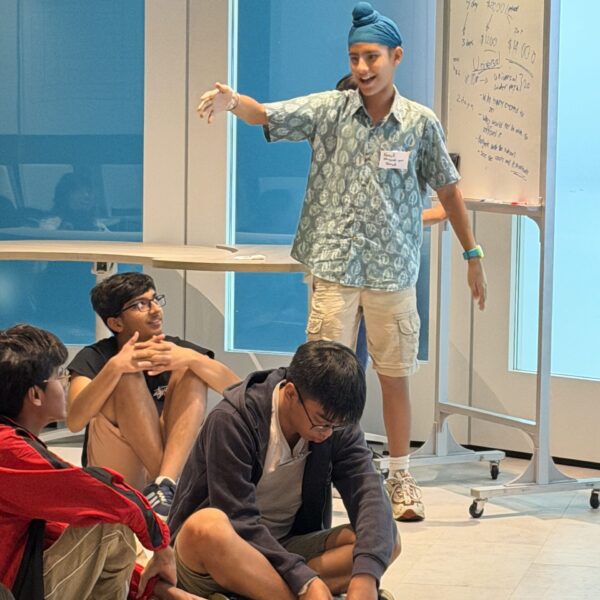 A young man in a blue turban gestures while presenting to a group of peers sitting on the floor at Youth Leadership and communication camp