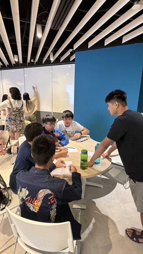 Students collaborating at a table in a modern study space with others writing on a whiteboard behind them