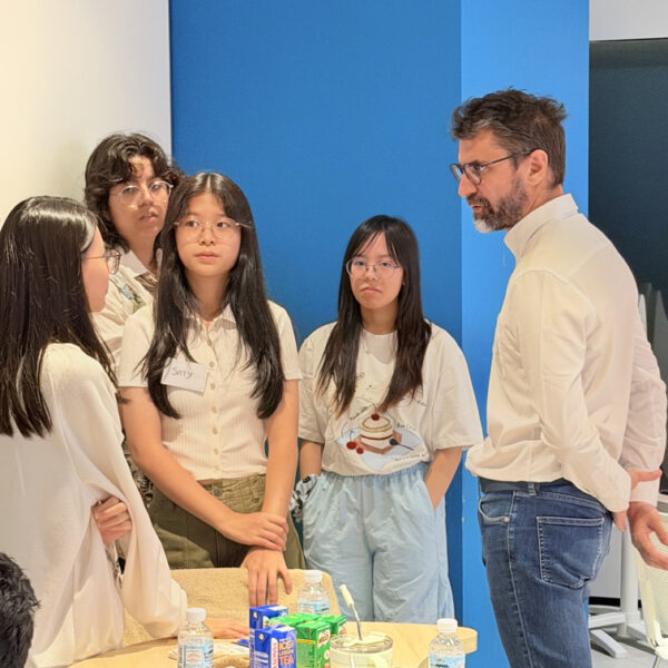 A man in a white shirt speaks to a group of four young students standing around a table with drinks