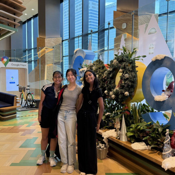 Three young women pose in front of a holiday-themed Google logo display in a bright office lobby