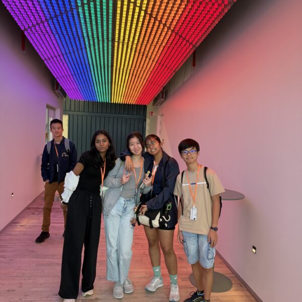 Five young people pose in a hallway beneath a vibrant rainbow light installation on the ceiling