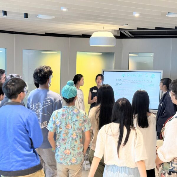 Two students present to a group of peers gathered around a digital whiteboard in a workshop setting