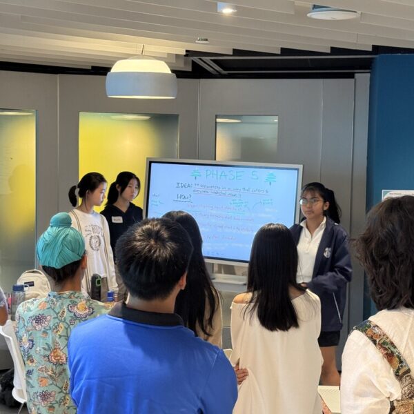 A student presents a project titled Phase 5 on a digital whiteboard to a group of peers while being filmed