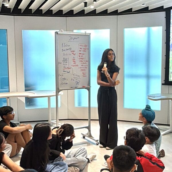 A young woman leads a workshop, standing by a whiteboard while a group of peers sit on the floor listening at Youth Leadership and Communication Camp
