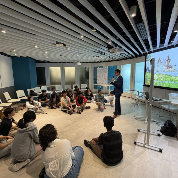 Students sit in a circle on the floor, listening to a speaker in front of a screen in Youth Leadership and Communication Camp