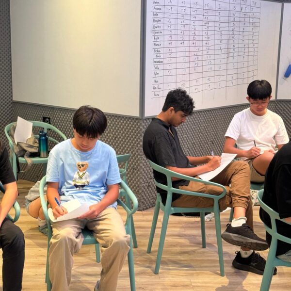 Three Youth Leadership and Communication Camp participants sit in chairs, writing on papers in front of a whiteboard